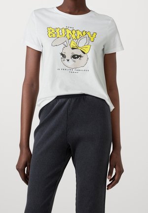 Biała bawełniana koszulka z grafiką królika w żółtej kokardzie. Połączona z ciemnymi, prążkowanymi spodniami, które mają luźny krój i fakturę.
