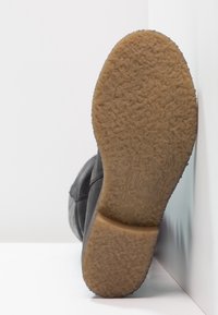 Bota de ante negra vista desde abajo, con una suela de goma texturizada en marrón claro que presenta un ligero patrón y un detalle de logo en relieve.