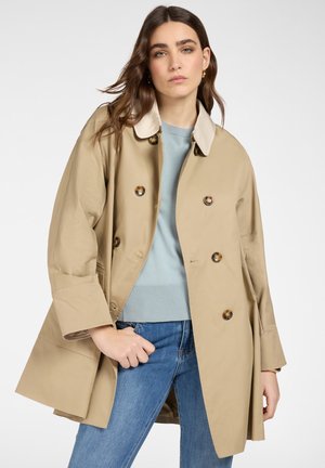 Giovane donna che indossa un trench doppiopetto beige sopra un maglione azzurro chiaro e jeans blu, in piedi contro uno sfondo neutro.