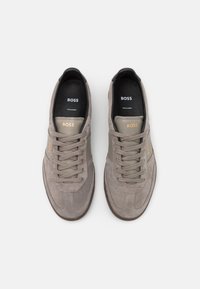 Šedé semišové tenisky mají zaoblenou špičku, tonální tkaničky a kontrastní béžový boční pruh. Na jazyku a boku je vyraženo logo "BOSS".