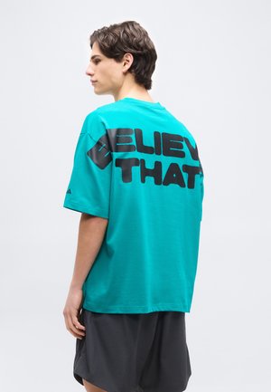 Junger Mann mit kurzen dunklen Haaren trägt ein übergroßes türkisfarbenes T-Shirt mit dem fetten schwarzen Schriftzug "BELIEVE THAT" auf dem Rücken und schwarze Shorts.