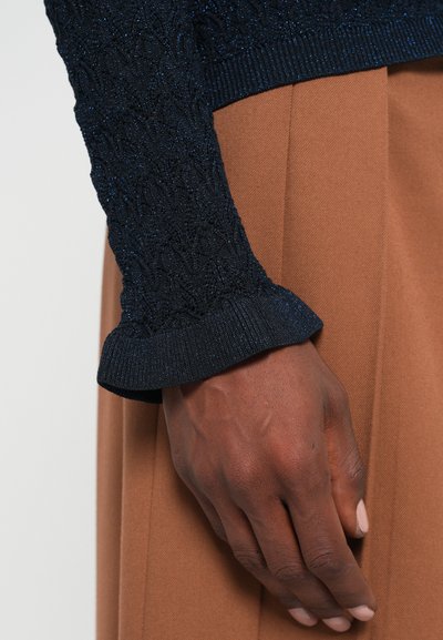 Pull en tricot bleu marine avec un motif texturé et des poignets côtelés, associé à un pantalon tailleur beige clair. Gros plan montrant les détails de la main et du tissu.