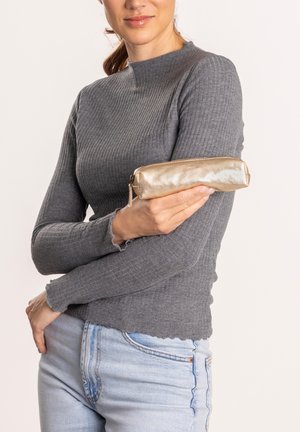 Metallic-goldener Federmäppchen mit glatter Oberfläche und runder Form, mit Reißverschluss, gehalten vor einem grauen gerippten Pullover und hellblauen Jeans.