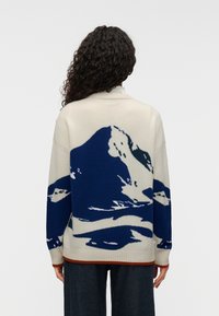 Πλεκτό πουλόβερ σε κρεμ χρώμα με μπλε σχέδιο βουνού. Διαθέτει ψηλό γιακά, μακριά μανίκια και ριμπ μανσέτες με μπορντό φινίρισμα.