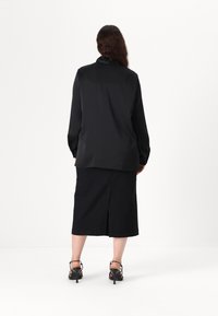 Black, long-sleeve blouse with a relaxed fit, paired with a knee-length black skirt featuring a back slit. Black strappy heels are worn.