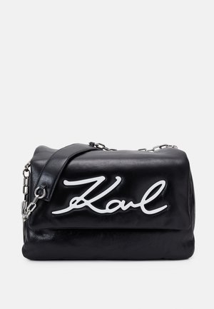 Czarna skórzana torba na ramię z srebrnym łańcuszkiem jako paskiem i białym kursywą napisem "Karl" na przedniej klapce.