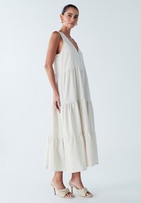 Kremowa sukienka maxi z głębokim dekoltem w serek, warstwowym wzorem tkaniny i detalami w postaci guzików. Noszona z plecionymi sandałami na obcasie z odkrytymi palcami.