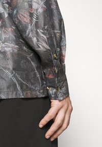Bluse mit floralem Muster aus leichtem Stoff in dunklen Tönen. Besitzt eine Knopfarme mit goldenen Akzenten und eine taillierte Form.