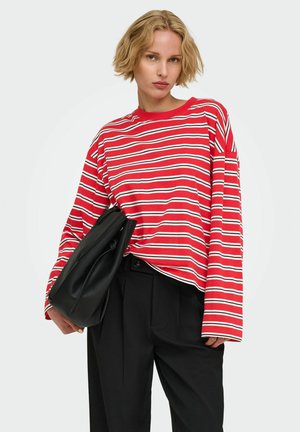 Kobieta z krótkimi blond włosami, ubrana w czerwoną i białą pasiastą bluzkę z długim rękawem oraz czarne spodnie, trzymająca pod pachą czarną torbę.