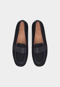 Ζευγάρι μαύρα πλεκτά μοκασίνια με δερμάτινες λεπτομέρειες και λουράκι, ταμπά εσωτερικές σόλες με την επωνυμία AGL, απεικονιζόμενα από πάνω σε λευκό φόντο.