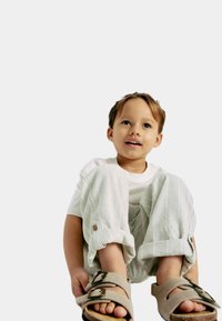 Kind dat een wit t-shirt en lichtgrijze gestreepte broek draagt, zittend met de voeten in beige sandalen met donkere gespen. Eenvoudige achtergrond.