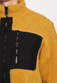 Giacca in fleece con un corpo strutturato giallo e accenti in nylon nero. Presenta un colletto alto, chiusura frontale con zip e tasca sul petto con dettagli rossi.