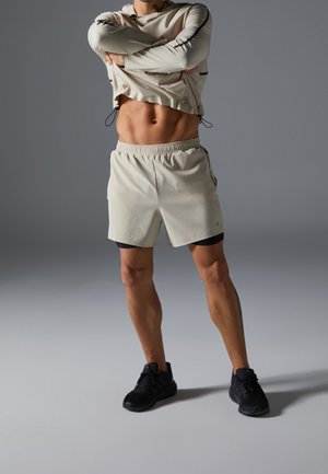 Athletischer Mann hebt beiges Langarmshirt, um seine definierten Bauchmuskeln zu zeigen, trägt beige Shorts und schwarze Laufschuhe vor grauem Hintergrund.