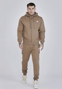 Sweat à capuche marron zippé avec cordons, arborant un logo sur la poitrine, assorti d'un pantalon de jogging marron avec poches latérales.