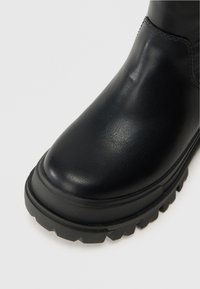 Fekete bőr bokacsizma lekerekített orral, texturált felülettel, vastag fekete gumi talppal, és rétegzett részletezéssel a varratok mentén.