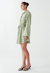 Šviesiai žalia suknelė su ilgomis rankovėmis, susiaurinta talija ir platėjančiu sijonu. Dėvima su juodomis šlepetėmis. Lygus audinys, laisvas pjūvis.