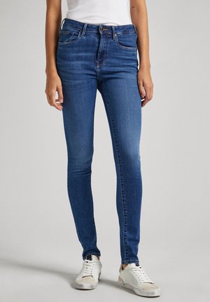 Vaqueros ajustados de denim azul con una cintura media, cinco bolsillos y un desgaste sutil. Combinados con zapatillas blancas. Textura suave.
