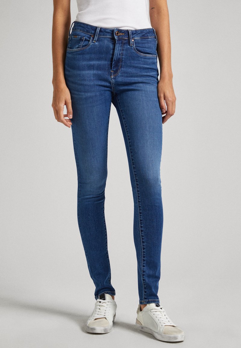 Vaqueros ajustados de denim azul con una cintura media, cinco bolsillos y un desgaste sutil. Combinados con zapatillas blancas. Textura suave.