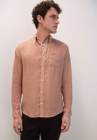 Camicia a maniche lunghe color ruggine con motivo geometrico a diamante, colletto abbottonato e taschino singolo sul petto. Tessuto liscio, design aderente.