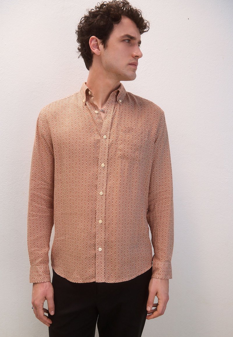 Camicia a maniche lunghe color ruggine con motivo geometrico a diamante, colletto abbottonato e taschino singolo sul petto. Tessuto liscio, design aderente.