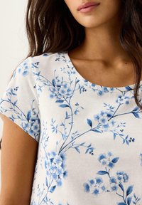 Kortærmet, rundhalset top i hvid bomuld med blå blomsterprint med stilke og blade, præsenterer en blød tekstur og detaljeret mønster.
