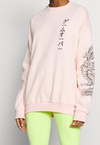 Sudadera rosa claro con texto en japonés, un diseño gráfico de dragón en la manga izquierda y puños y dobladillo acanalados. Combinada con leggins verde neón.