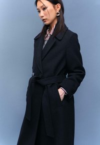 Čierny vlněný kabát s priliehavým strihom, s opaskom v páse, veľkým golierom a zapínaním na gombíky. Model má pod ním pruhované tričko.