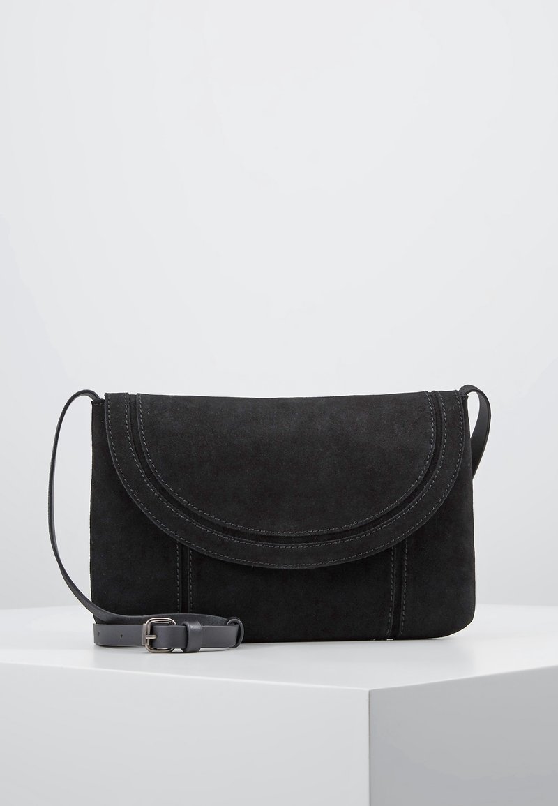 Černá semišová taška přes rameno s zakřivenou klopou a nastavitelním koženým popruhem, vystavená na bílé podstavě na jednoduchém světlém pozadí.