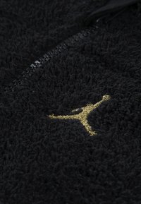 Fekete textúrájú anyag, bolyhos megjelenéssel, kiemelkedő arany hímzett Jumpman logóval és fekete cipzár részlettel.