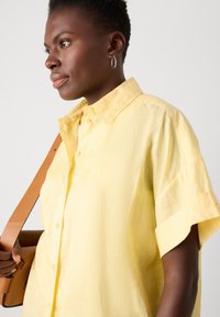 Camicia gialla con bottoni, colletto e maniche corte, caratterizzata da un tessuto strutturato. Dettaglio significativo include la cucitura sulla manica.