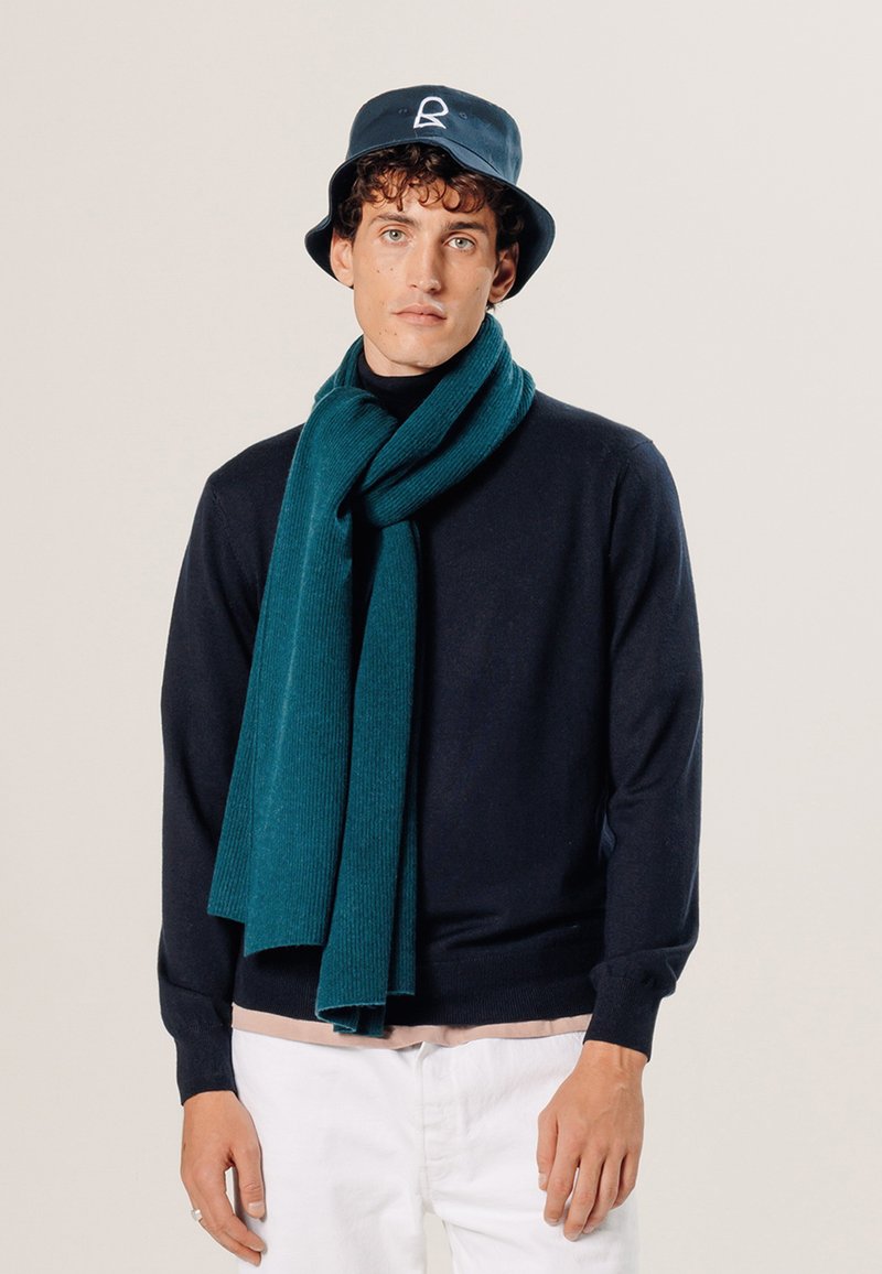 Marineblauer Pullover mit einem gerippten türkisfarbenen Schal. Das Model trägt einen marineblauen Bucket Hat mit Logo. Der Hintergrund ist neutral. Auffällige Texturen: weich und gestrickt.