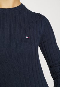 Pull en maille côtelée bleu marine à manches longues avec un petit logo rectangulaire rouge, blanc et bleu sur la poitrine gauche.