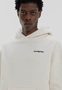Κρέμα χρώματος hoodie από μαλακό ύφασμα, με μπροστινή τσέπη και μαύρο λογότυπο "lowlights" στο στήθος. Χαλαρή εφαρμογή και κουκούλα με κορδόνι.