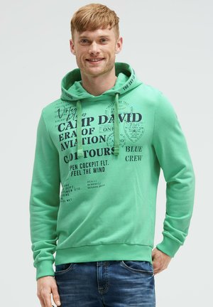 Lächelnder Mann, der einen grünen Hoodie mit schwarzem Luftfahrttext und blauen Jeans trägt, steht vor einem einfarbigen hellen Hintergrund.