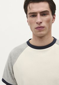 T-shirt en coton crème et gris avec des manches raglan courtes, un col bleu foncé et une texture lisse. Gros plan sur le haut du corps.