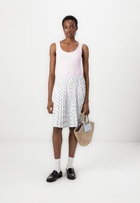 Débardeur rose pâle, associé à une jupe blanche ornée d'un motif géométrique noir. Sac tissé à la main, chaussures noires et chaussettes blanches.