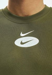 Žalia alyvuogių spalvos medvilninė marškinė su tekstūruotu apykaklės įpjovimu, ant kurių yra balta siuvinėjama Nike logotipas ir stilizuota svorio linija.