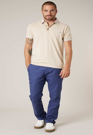 Homme avec des tatouages portant un polo beige, un pantalon bleu et des baskets blanches, debout avec une main dans la poche sur un fond uni.