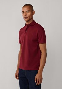 Burgundská polo košeľa s krátkymi rukávmi, golierom a jedným gombíkom pri krku. Vyrobená z hladkej látky, v kombinácii s tmavými denimovými džínsami.