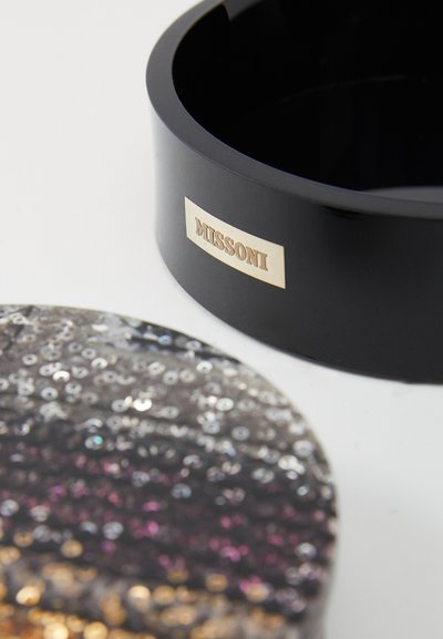 Μαύρος στρογγυλός πυθμένας με μεταλλικό λογότυπο Missoni, συνοδευόμενος από καπάκι με μοτίβο πολύχρωμων παγιέτας σε γραδαριστές αποχρώσεις.