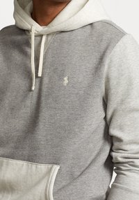 Grå hoodie med en kontrasterande ljusgrå ficka och inre foder. Har en huva med dragsko och en liten broderad logga på bröstet.