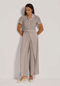 Vrouw met lang zwart haar, gekleed in een taupe blouse met korte mouwen en kraag, en wijde broek met knoopdetail, beige slingback hakken, staande tegen een witte muur.