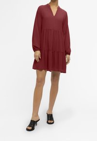 Robe rouge jusqu'aux genoux avec manches longues, dotée d'un col en V et d'un design à étages. Portée avec des mules à talons noires ouvertes à l'avant.