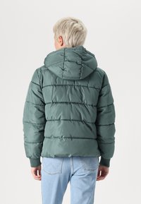 Zelena pufasta jakna s kapuljačom, koja ima horizontalni quilted dizajn, elastične manžete i glatku, sjajnu teksturu. Modernog je, ležernog kroja.