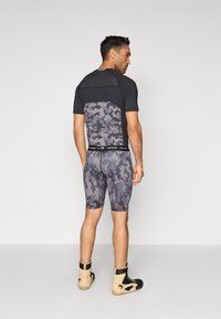 Camisola preta desportiva ajustada com padrão camuflado cinzento nas costas, combinada com calções camuflados iguais e calçado de cor clara.