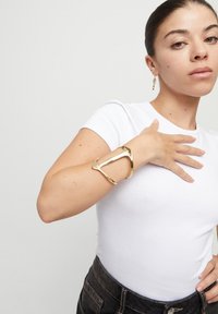 Pulsera de metal dorado en forma de brazalete con un diseño triangular abierto, que presenta superficies lisas y texturizadas, llevada en la muñeca de un modelo.