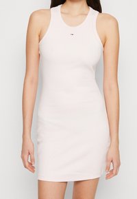 Žena v přiléhavých, bezrukávových, světle růžových mini šatech s malým logem na hrudi na jednobarevném pozadí.