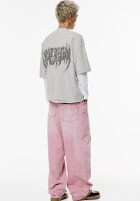 T-shirt grigio oversize con una grafica testurizzata sulla schiena; abbinato a jeans larghi color rosa pastello. Il modello indossa scarpe nere e abbigliamento a strati.