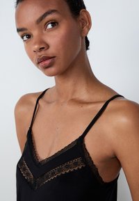 Camisola nera con dettagli in pizzo, caratterizzata da spalline sottili regolabili e scollo a V, mostrata su uno sfondo neutro.