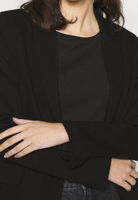 Czarna marynarka z kołnierzem szalowym, teksturowana tkanina, szerokie rękawy i subtelne detale kieszeni, zestawiona z dopasowanym czarnym prążkowanym topem pod spodem.